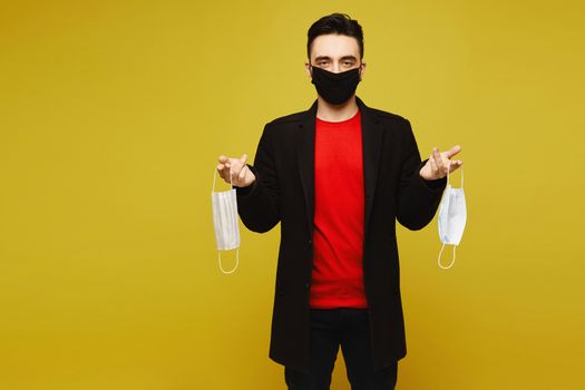 Young man in a protective medical mask holding a pair of protective masks in hands, isolated in a yellow background. Medical advertisement mockup.