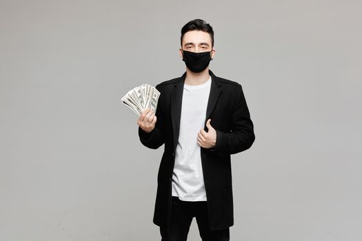 Handsome young man in black protective mask holding a pack of money, isolated on grey background with copy space. Concept of medical and pharmaceutical business in a season of flu and world pandemic. Men wearing medical mask to prevent infection, airborne respiratory illness such as flu, 2019-nCoV.