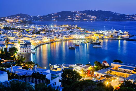 View of Mykonos Chora town Greek tourist holiday vacation destination with famous windmills, and port with boats and yachts illuminated in the evening blue hour . Mykonos, Cyclades islands, Greece