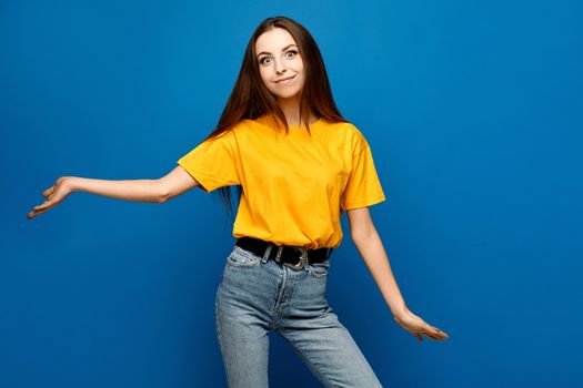 A young surprised woman in yellow blank shirt stands with arms apart over blue background, isolated.