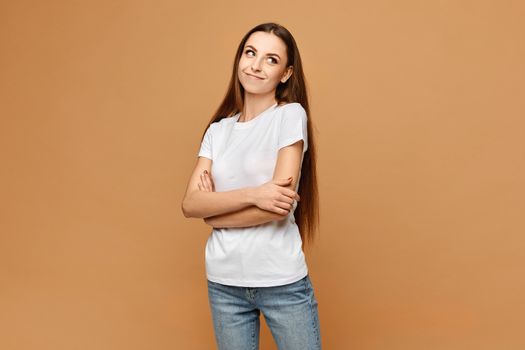 Young woman in white blank t-shirt and blue jeans posing with folded hands at beige background, isolated. Model girl in a casual outfit. Mockup for advertising.