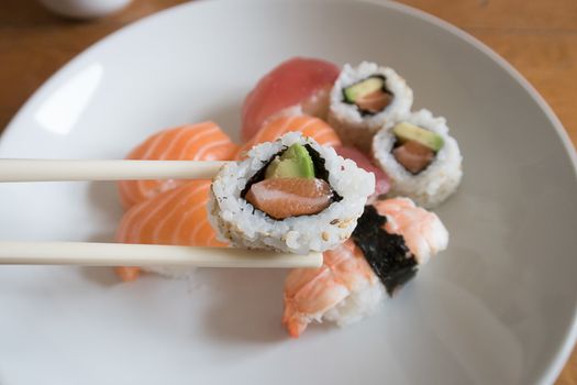 Fresh salmon and avocado uramaki sushi between chopsticks with salmon nigiri, prawn nigiri and tuna nigiri in the background