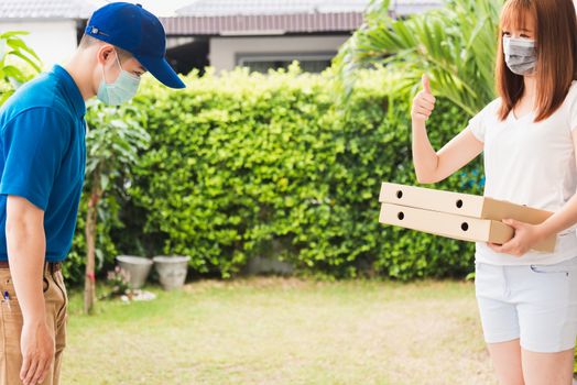 Asian delivery courier young man giving food pizza box to woman customer receive both protective face mask and show thumb up finger for good job, under curfew quarantine pandemic coronavirus COVID-19