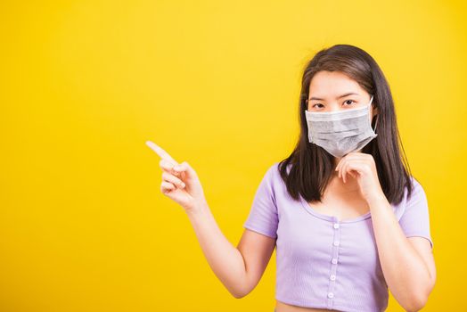 Asian young teen woman wearing face mask protective against coronavirus or COVID-19 virus or filter dust, air pollution her point finger to side space, studio shot isolated yellow background