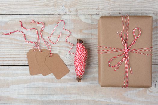 Overhead view of blank gift tags a spool of string and a plain brown paper wrapped present on a rustic whitewashed wood table. 