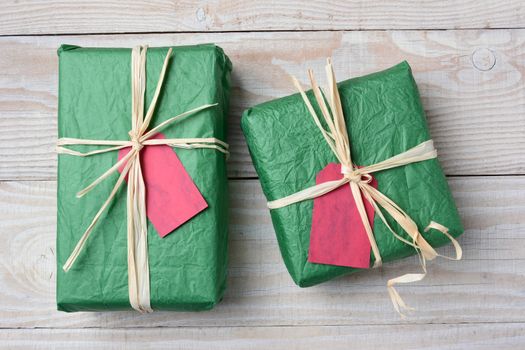 High angle image of two green Christmas presents on a rustic white wood table. Wrapped with crumpled tissue paper and tied with raffia both have blank red gift tags. Horizontal format.