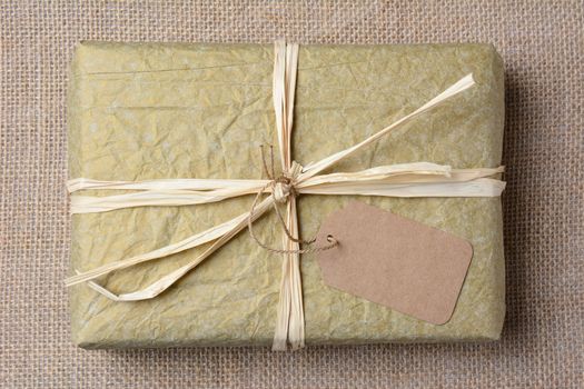 Closeup of a gold tissue paper wrapped present on a burlap surface. The gift is tied with raffia and a blank git tag. High angle shot in horizontal format. 