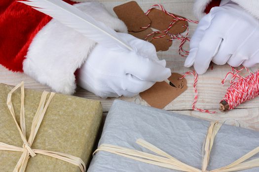 Santa Claus sitting at his desk writing gift tags for Christmas presents. Horizontal closeup showing gifts and Santa's hands only.