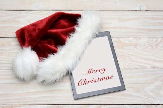 High angle shot of a tablet computer with a Santa Claus hat over one corner. The screen is white with the words Merry Christmas in red type. Type is easily removed to add you own message.