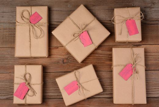 Six holiday packages wrapped in brown kraft paper and tied with twine. The parcels are all different sizes and shapes and resting on a rustic wooden table. There is a blank gift tag attached to each gift. Shot form a high angle.