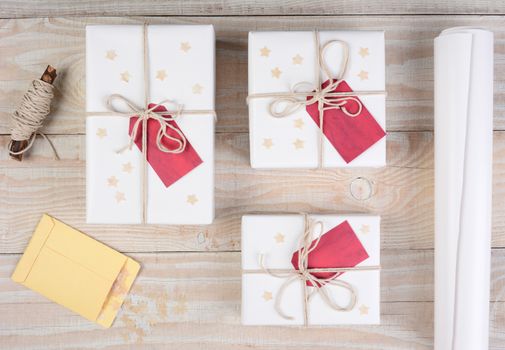 High angle shot of Christmas presents wrapped in white paper and tied with white string. Red gift tags and stars adorn the packages on a white wood table.