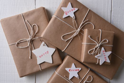 A pile of Christmas presents wrapped with plain brown paper, tied with string and with star shaped tags stamped with the word Merry.