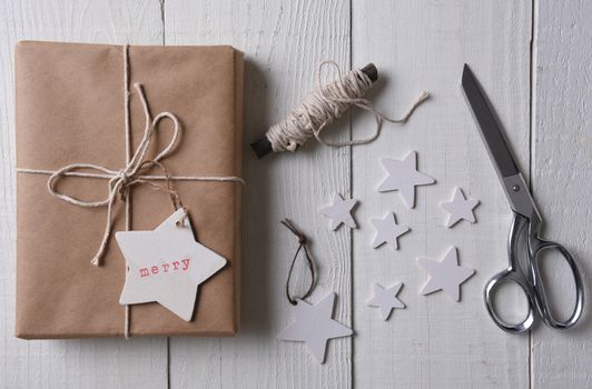 Top view of a single plain paper wrapped Christmas present with a tag stamped with Merry. Extra stars, twine and scissors are next to the gift.