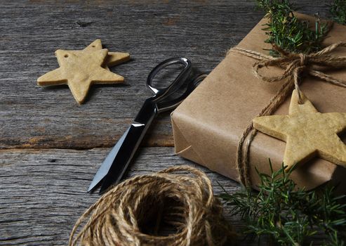 Christmas present wrapped in plain brown paper on a rustic wood table with holiday cookies, twine, scissors and greenery.
