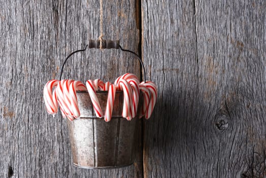 Christmas Candy Canes: A bucket full of the peppermint holiday treat hanging from twine against a rustic wood wall.