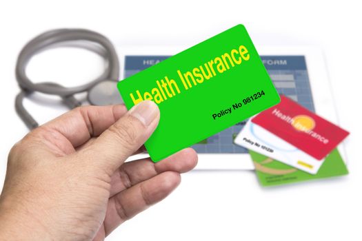 Hand holding green health insurance card in front of tablet and other insurance card on white background.