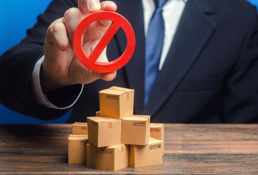 Businessman man official holding red prohibition sign NO over boxes with goods. Out of stock. Embargo trade wars. Restriction import, ban export of dual use goods to countries under sanctions.