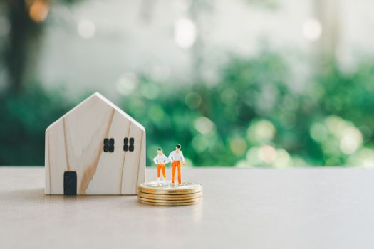 Saving money concept. Miniature people or small model businessman figure standing on coins stack with wooden home on green nature background.