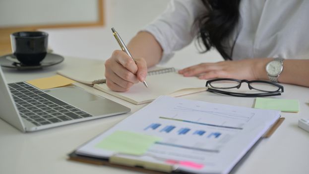 close-up shot of Girl with a pen in hand is recording the company performance with graph and laptop on the desk.