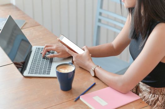 Cropped shot of a woman holding a smartphone and using a laptop on a table with a coffee cup in a modern workplace.