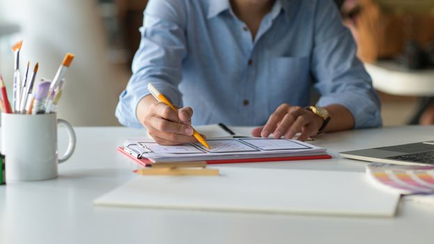 Cropped shot of Designer is sketching a smartphone screen for future customers on the desk with laptop and stationeries.