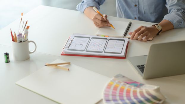 Cropped shot of young designer working on smartphone application project with tablet and notebook in modern office room.