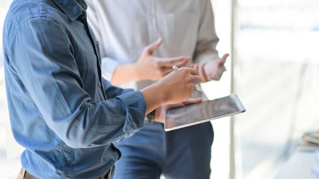 Two young businessmen consulted about their work by using a tablet while standing at working place, Side view image.