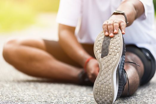 Close up Asian young athlete sport runner black man wear watch he sitting pull toe feet stretching legs and knee before running at outdoor street health park, healthy exercise before workout concept