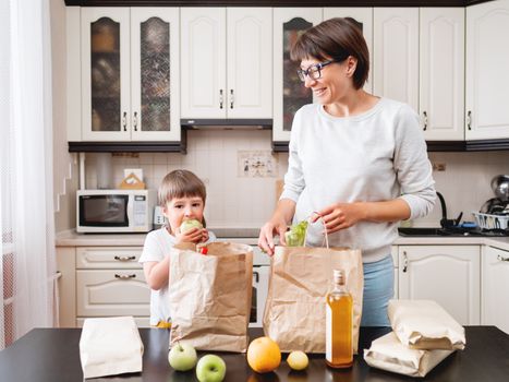 Woman and toddler sorts out purchases in the kitchen. Kid bites an apple. Grocery delivery in paper bags. Subscription service from grocery store. Mother and son at kitchen.