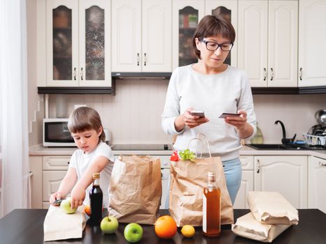 Woman and toddler boys sorts out purchases in the kitchen. Kid bites an apple. Grocery delivery in paper bags. Subscription service from grocery store. Mother and son at kitchen.