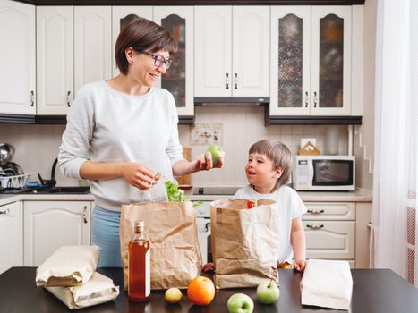 Woman and toddler boy sorts out purchases in the kitchen. Kid bites an apple. Grocery delivery in paper bags. Subscription service from grocery store. Mother and son at kitchen.