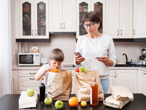 Woman and toddler boy sorts out purchases in the kitchen. Grocery delivery in paper bags. Subscription service from grocery store. Mother and son at kitchen.