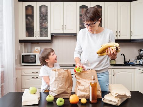 Woman and toddler boy sorts out purchases in the kitchen. Kid bites an apple. Grocery delivery in paper bags. Subscription service from grocery store. Mother and son at kitchen.