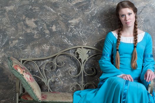 Pretty young woman in historical medieval blue dress sits on a banquette in studio