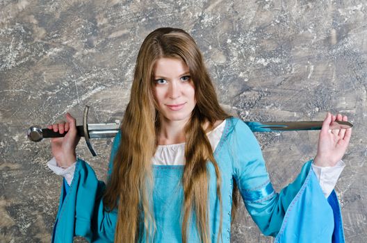 Pretty young woman with long hair in historical medieval blue dress poses in studio with sword