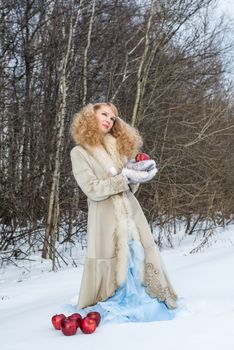 Young woman in a fur coat poses at winter forest with apples