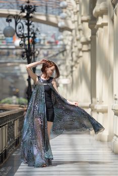 Pretty tender young model is walking in a short black dress with fashionable flutter transparent cape at old fashioned building with pillars