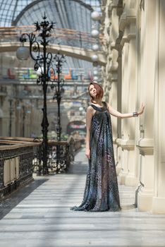 Caucasian tender young model is posing in a short black dress and lengthy transparent cape at old fashioned building with pillars