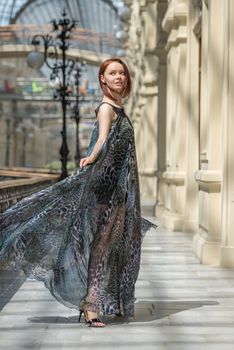 Pretty tender young model is walking in a short black dress with fashionable flutter transparent cape at old fashioned building with pillars