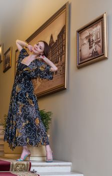 Charming sensual young woman in gauzy lengthy dress is posing at historic ladder near paintings on a wall