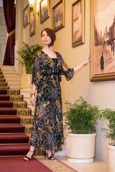 Charming sensual young woman in gauzy lengthy dress is posing at historic ladder near paintings on a wall