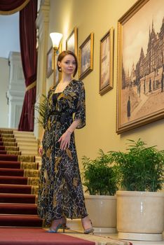 Charming sensual young woman in gauzy lengthy dress is posing at historic ladder near paintings on a wall