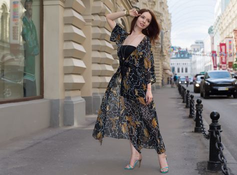 Charming sensual woman in fashionable gauzy clothing is posing at a town street with cars and people at blurred background