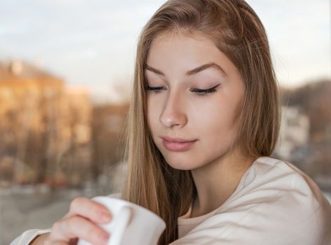 Lovely girl is sitting indoor near the window and enjoying the aroma from a white mug