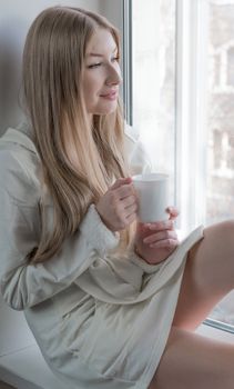 A happy young girl in a short dressing gown is sitting with mug near the window