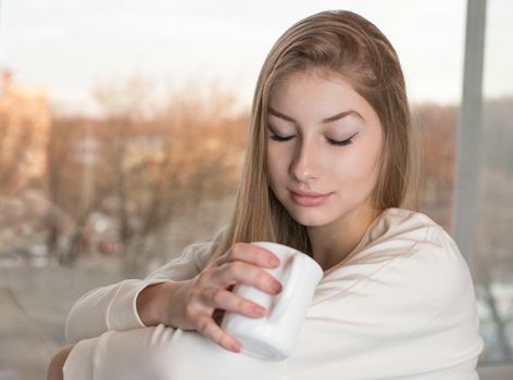 Charming dreamy girl is sitting indoor near the window and enjoying the aroma from a white mug