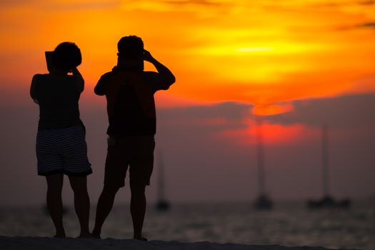 A silhouette of couple people are taking photo of sunset on the beach under colorful sky located at south of thailand