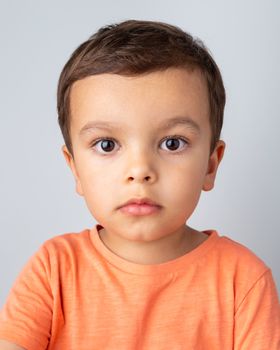 Cute three year old boy portrait, toddler wearing orange tee shirt and shot against a light grey background.