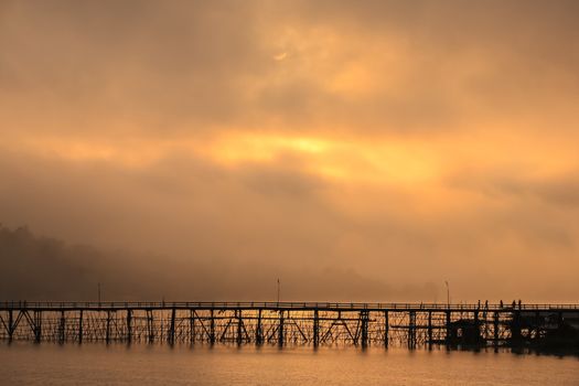 Wooden bridge across through river with foggy background at sunrise reflection on water located Kanchanaburee province west of Thailand