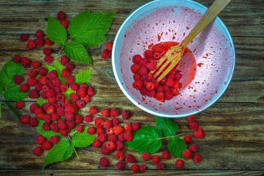 Red fresh raspberries on brown rustic wood background. Bowl with natural ripe organic berries with peduncles, green leaves and wooden fork, top view, flat lay with copy space.